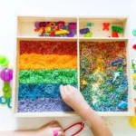 boîte-sensorielle-avec-riz-arc-en-ciel-à-l-intérieur-bébé-matériel-montessori-les-mains-des-enfants-touchent-la-texture-et-163453809